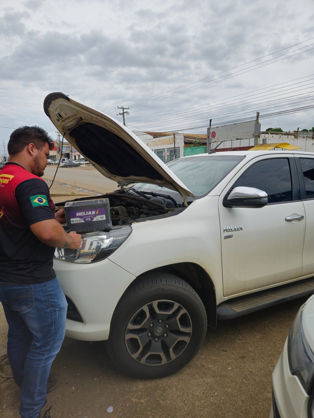 A mechanic installing a new Heliar car battery into a white Toyota Hilux truck with the hood open.
