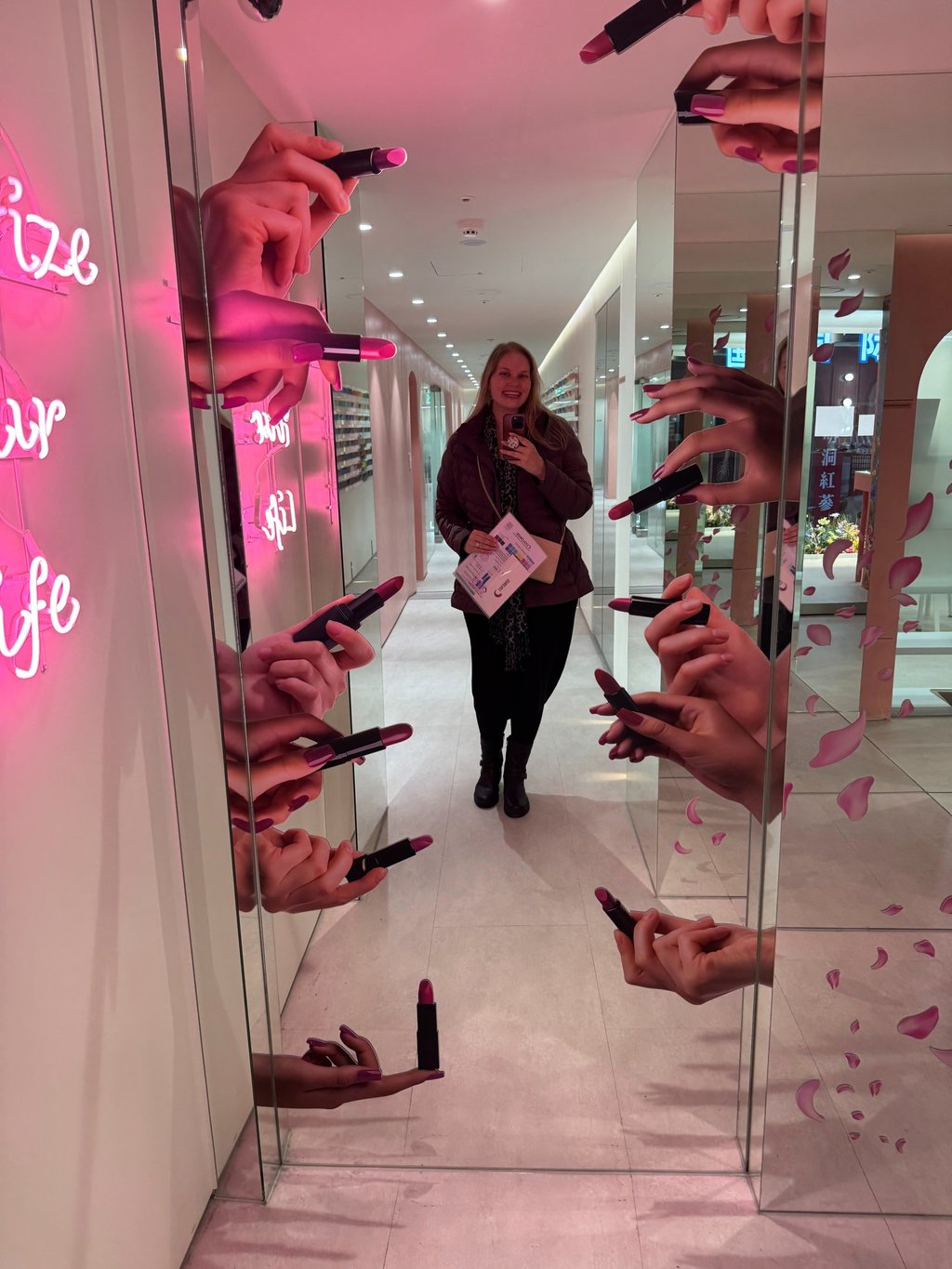 A woman takes a mirror selfie in a creative makeup display corridor with 3D hands holding lipsticks.