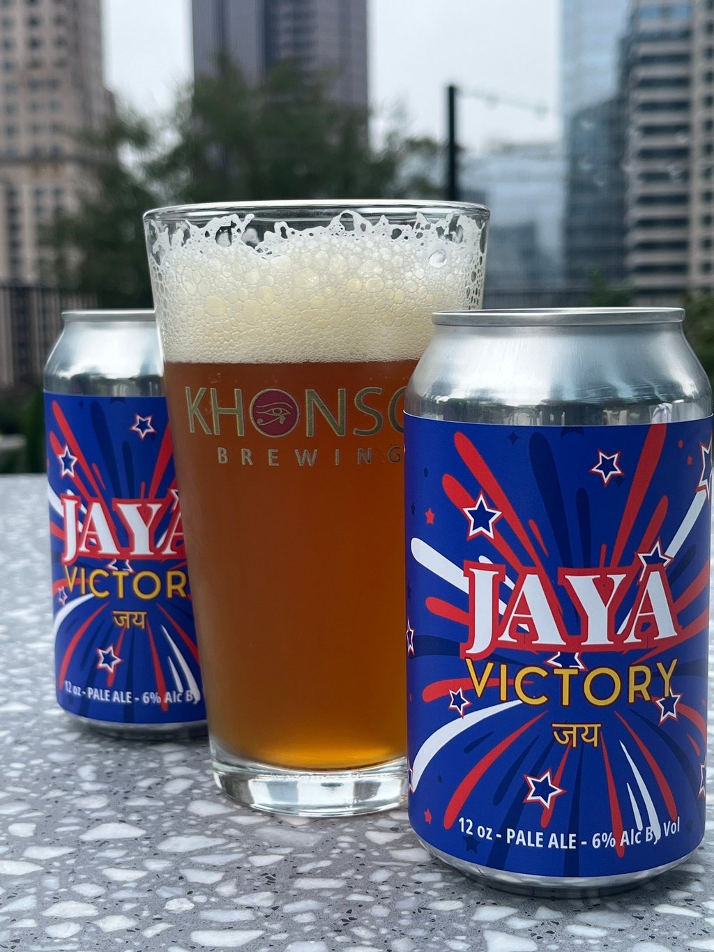 A glass of foamy Khonso Brewing pale ale next to Jaya Victory beer cans on a rooftop.