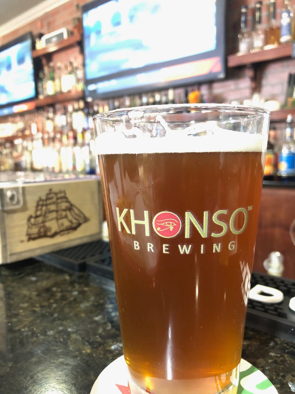 A pint of amber craft beer from Khonso Brewing sits on a bar counter with a blurred tavern background.