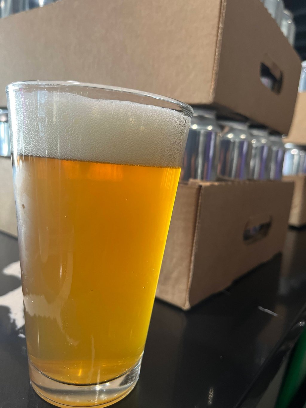 A cold pint of golden craft beer with foam head next to cardboard cases of canned beverages.