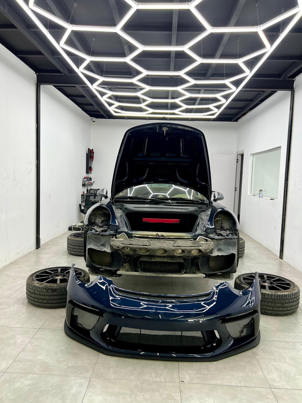 A dark blue sports car undergoing professional detailing and repair under hexagonal LED garage lights.