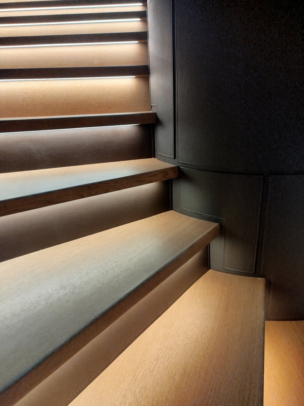 Modern wooden staircase with integrated LED tread lighting and dark leather wall panels.