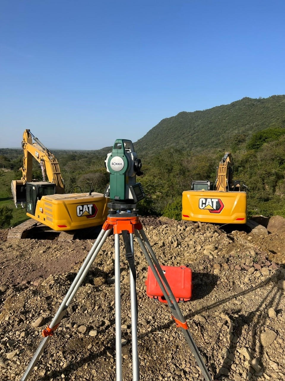 Sokkia total station on a tripod at a construction site with two CAT excavators and hills.