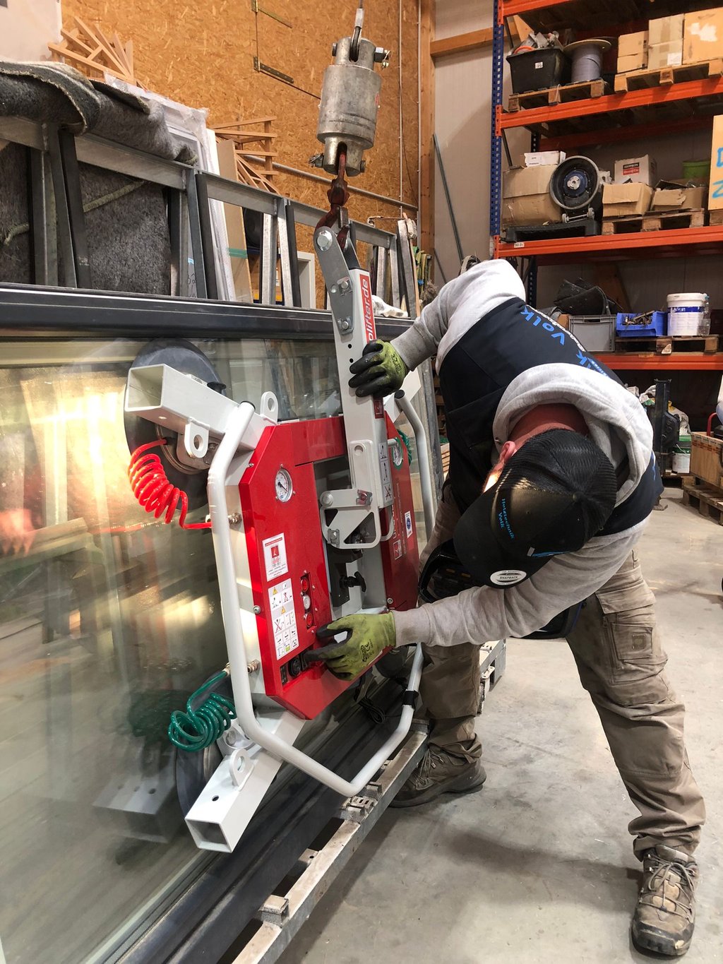 Schreinermeister Ferdinand Volk justiert den roten Vakuum-Saugheber an einer großen Glasscheibe, die an einem Kranhaken ist.