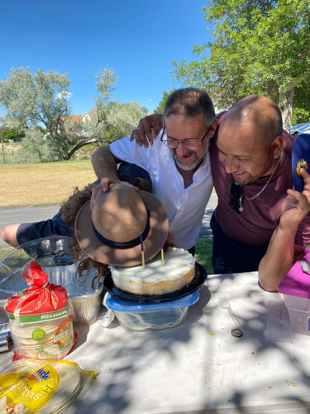 Friends gathered outdoors for a birthday celebration with a frosted cheesecake and candles.