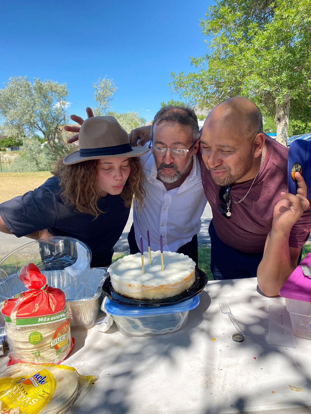 Three people blowing out purple candles on a round birthday cake at an outdoor family gathering.