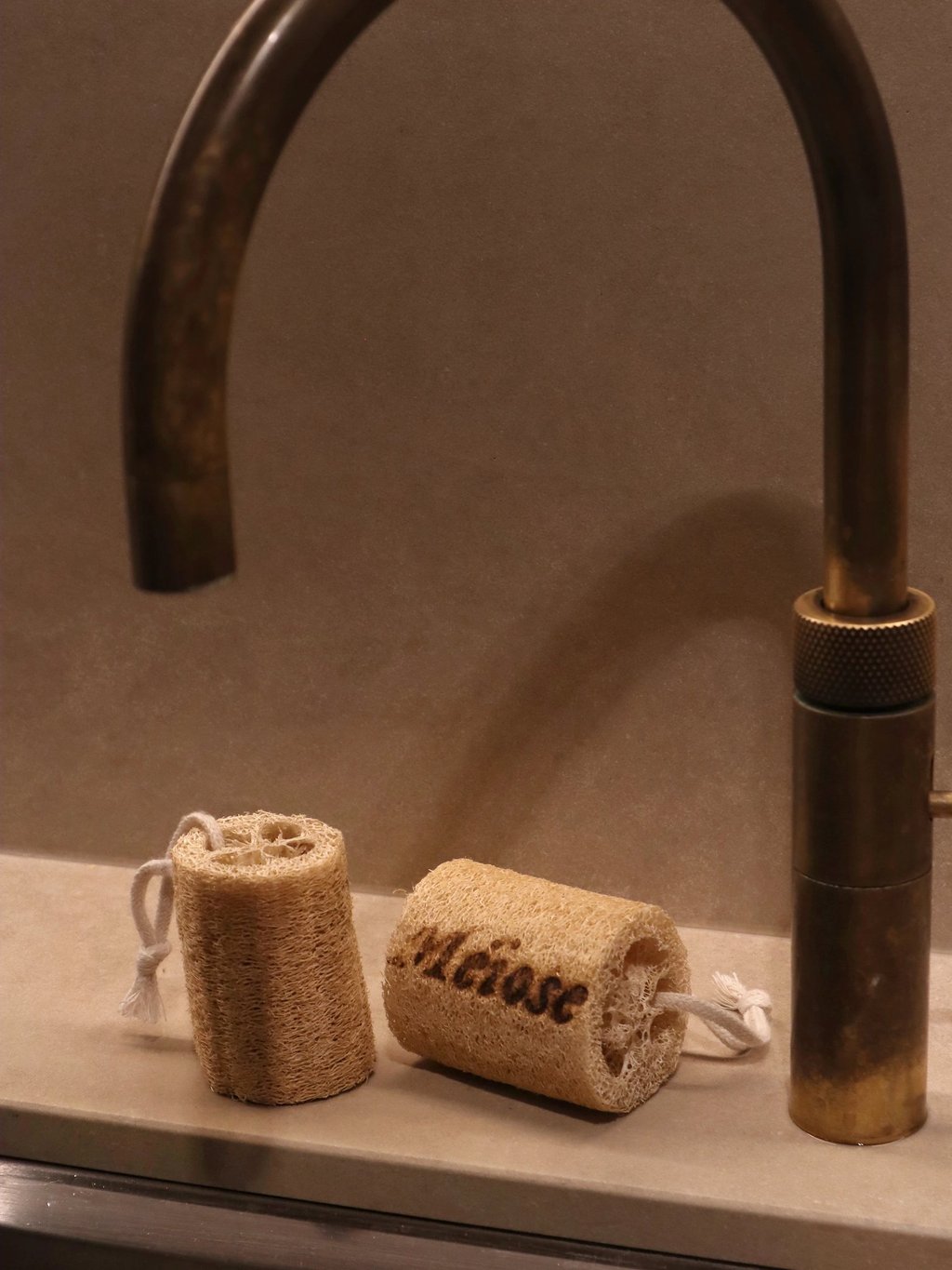Two natural loofah sponges with cotton loops sit on a stone counter next to a vintage brass faucet.