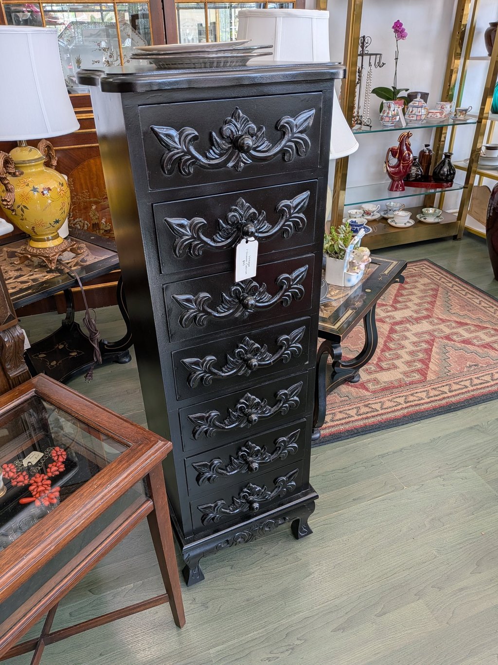 Elegant black lingerie chest with ornate floral wood carvings on seven drawers in an antique shop.