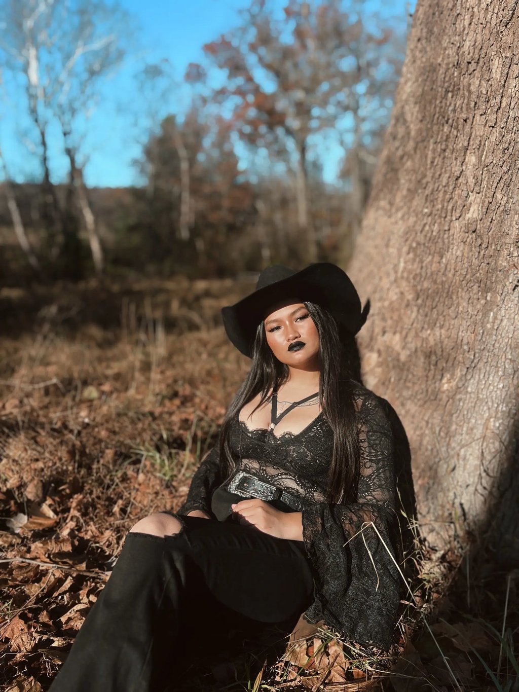 Amelia leaning against a tree in a black lace top with a black cowboy hat