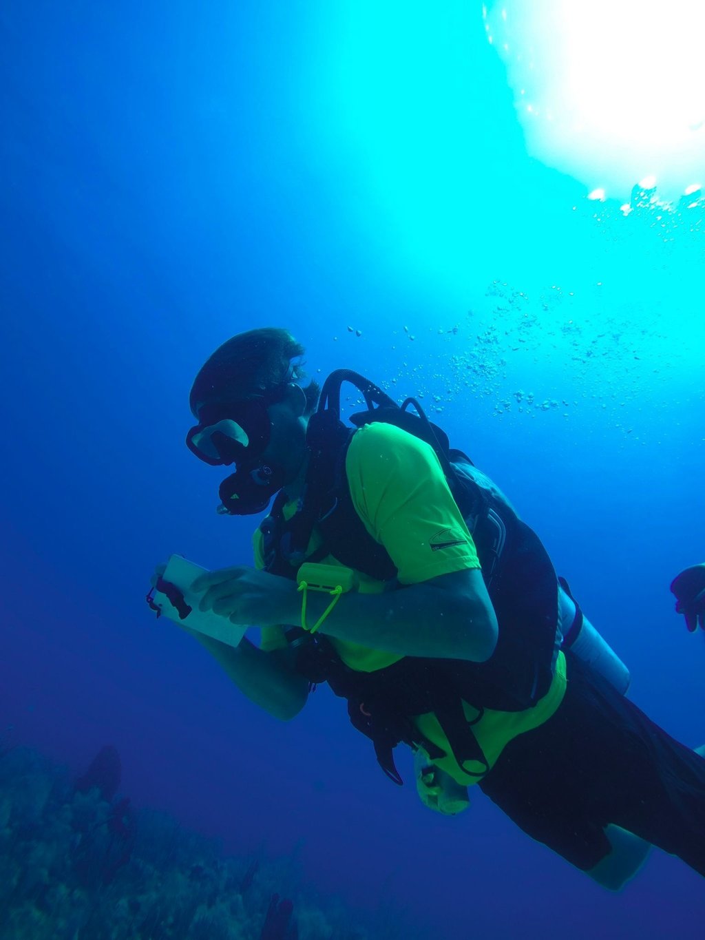 A scuba diver in yellow gear writes on an underwater slate while diving in deep blue ocean water.