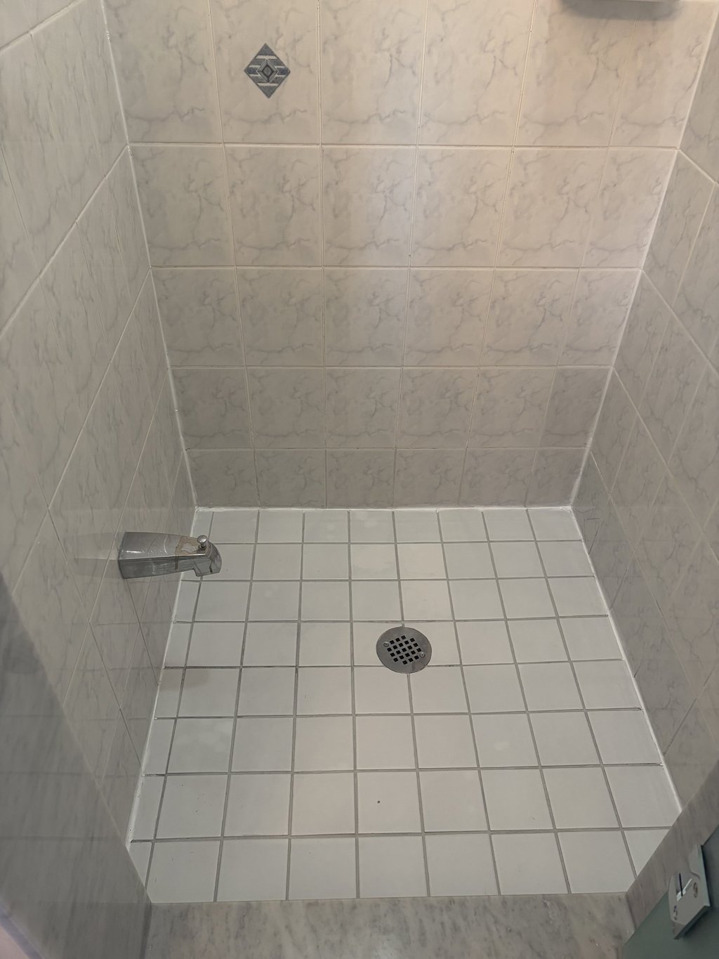 A walk-in shower featuring white floor tiles, a chrome drain, marble-patterned wall tiles, and a bathtub spout.