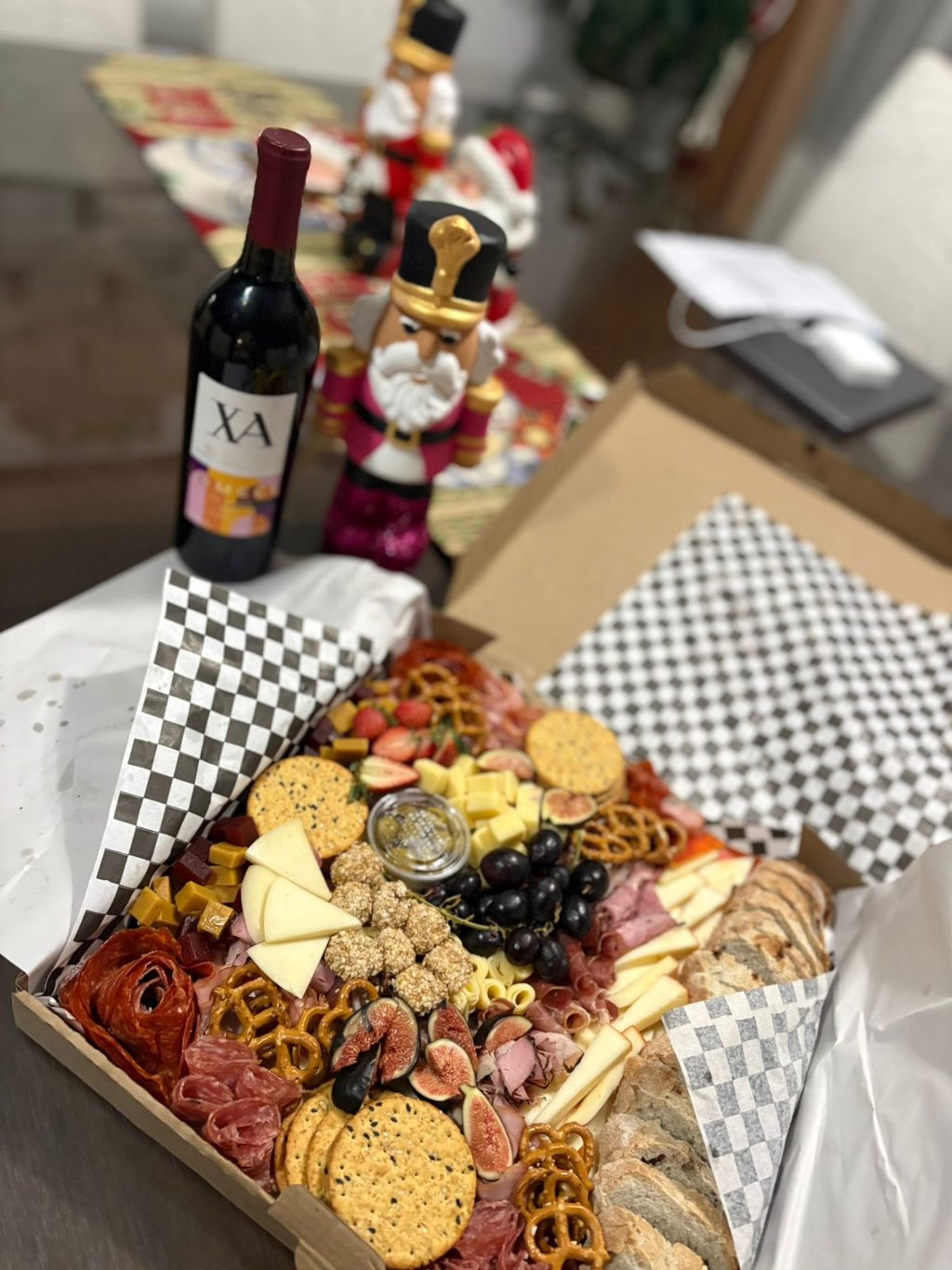 Gourmet charcuterie board box with cheeses, meats, and fruit next to wine and a Christmas nutcracker.