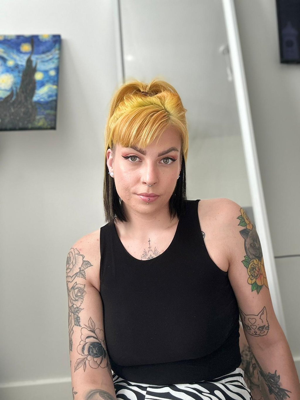 A woman with yellow and black split-dye hair and arm tattoos wearing a black tank top.