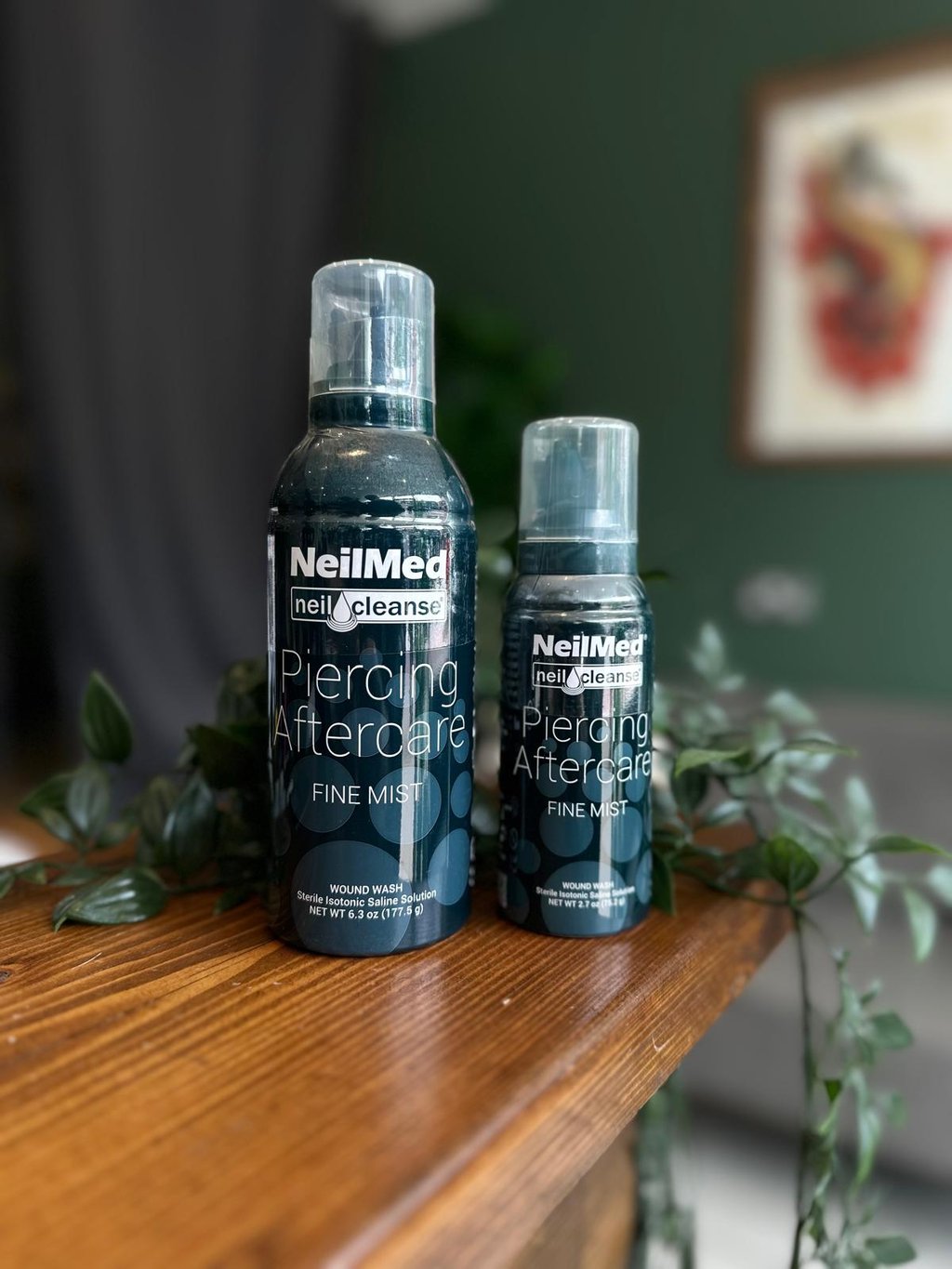 Two sizes of NeilMed Piercing Aftercare fine mist sterile saline solution spray on a wooden table.