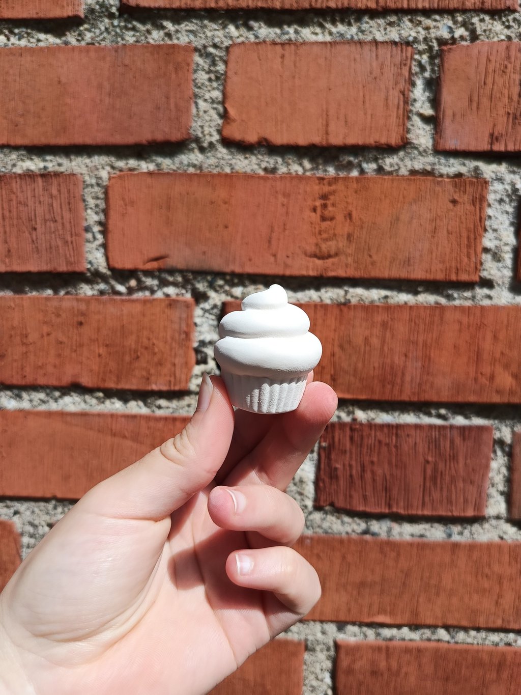 Category tags and tiny objects: Small white ceramic cupcake figurine held against a red brick wall.