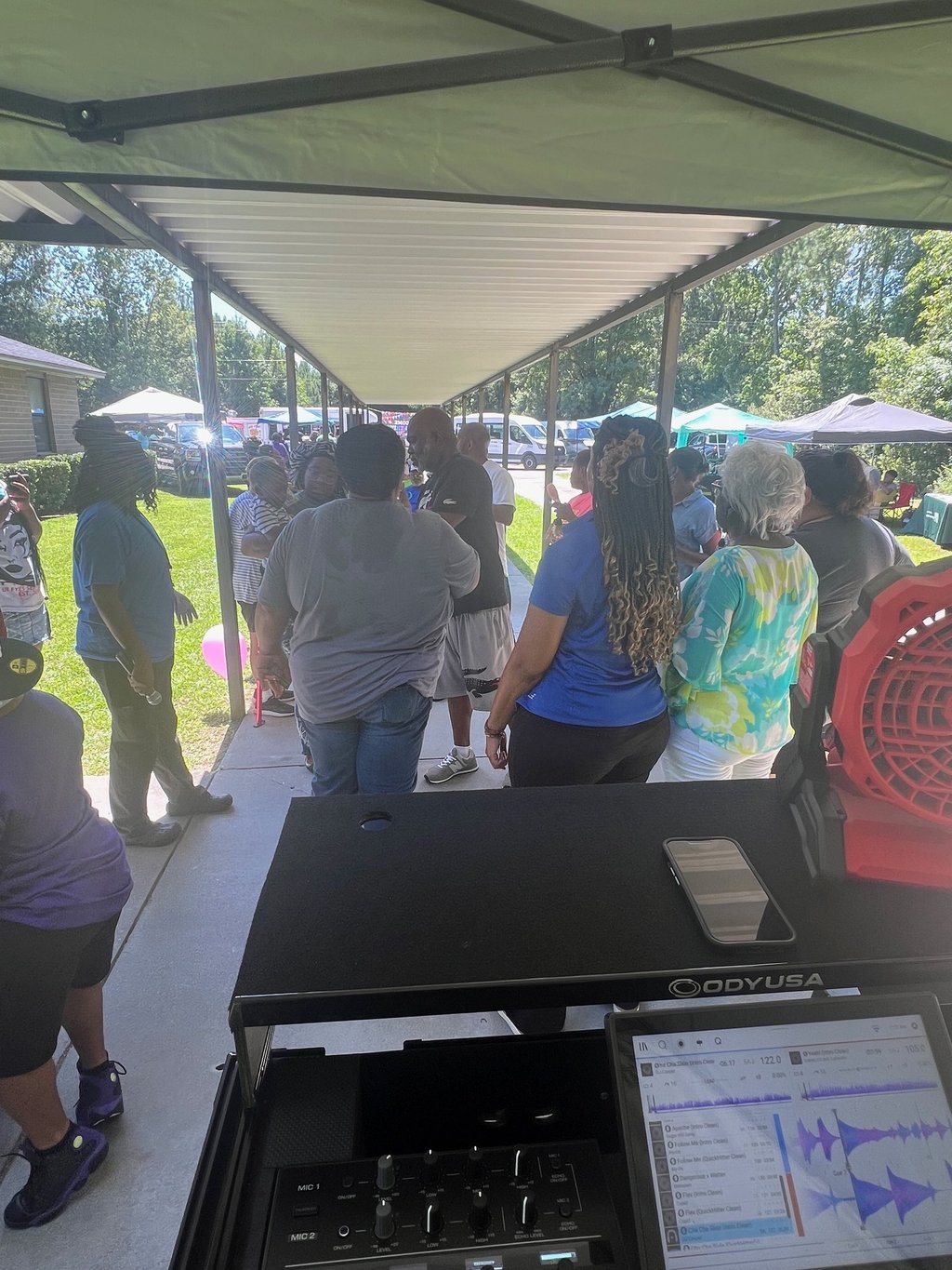 A DJ booth setup at an outdoor community event with people gathered under a long canopy.