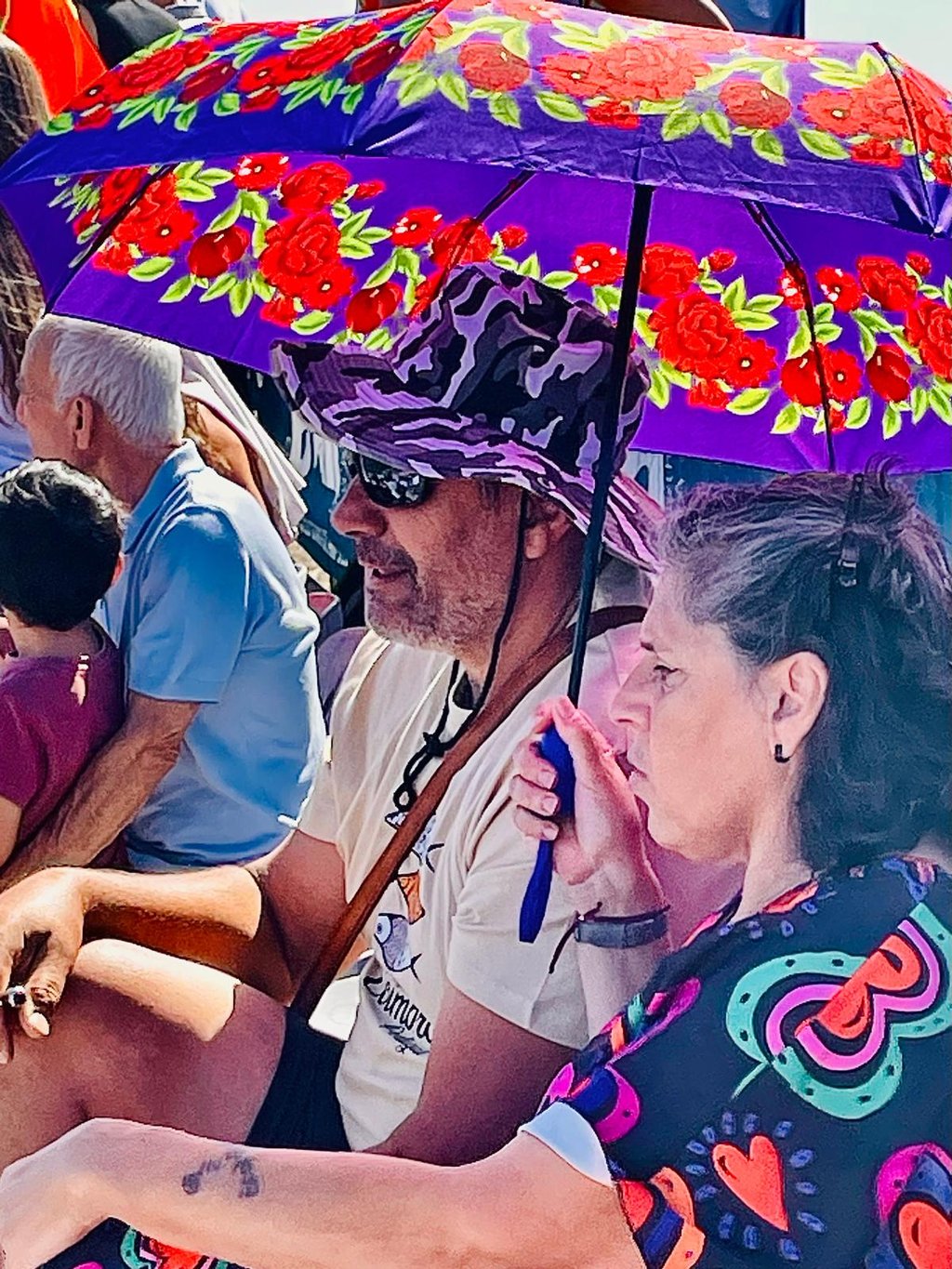 A man in a purple hat and a woman sit under a floral purple sun umbrella at an outdoor event.