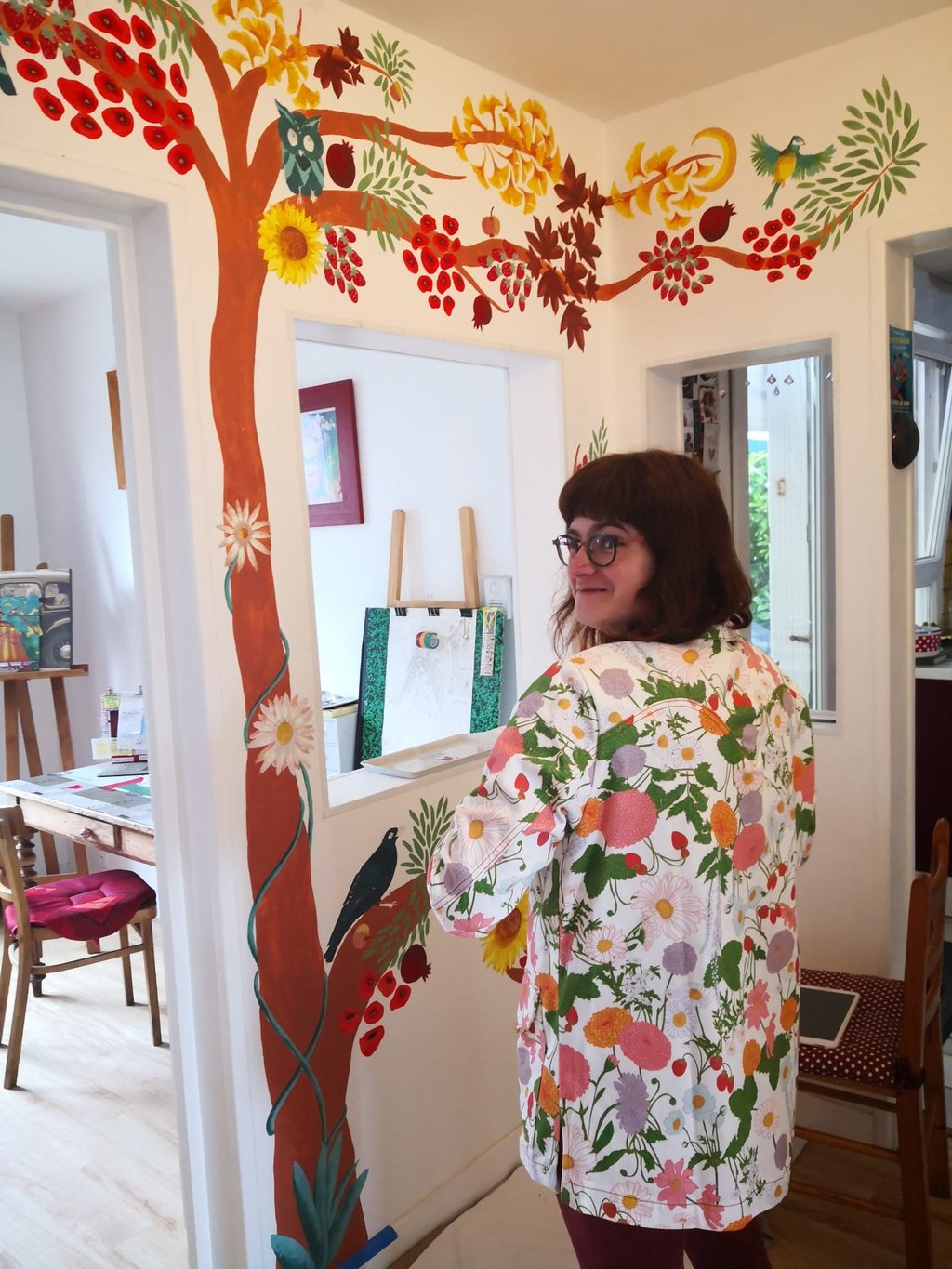 Photo de l'artiste-peintre Marie Hardy devant une fresque d'arbre réalisée dans le salon d'un particulier en Normandie