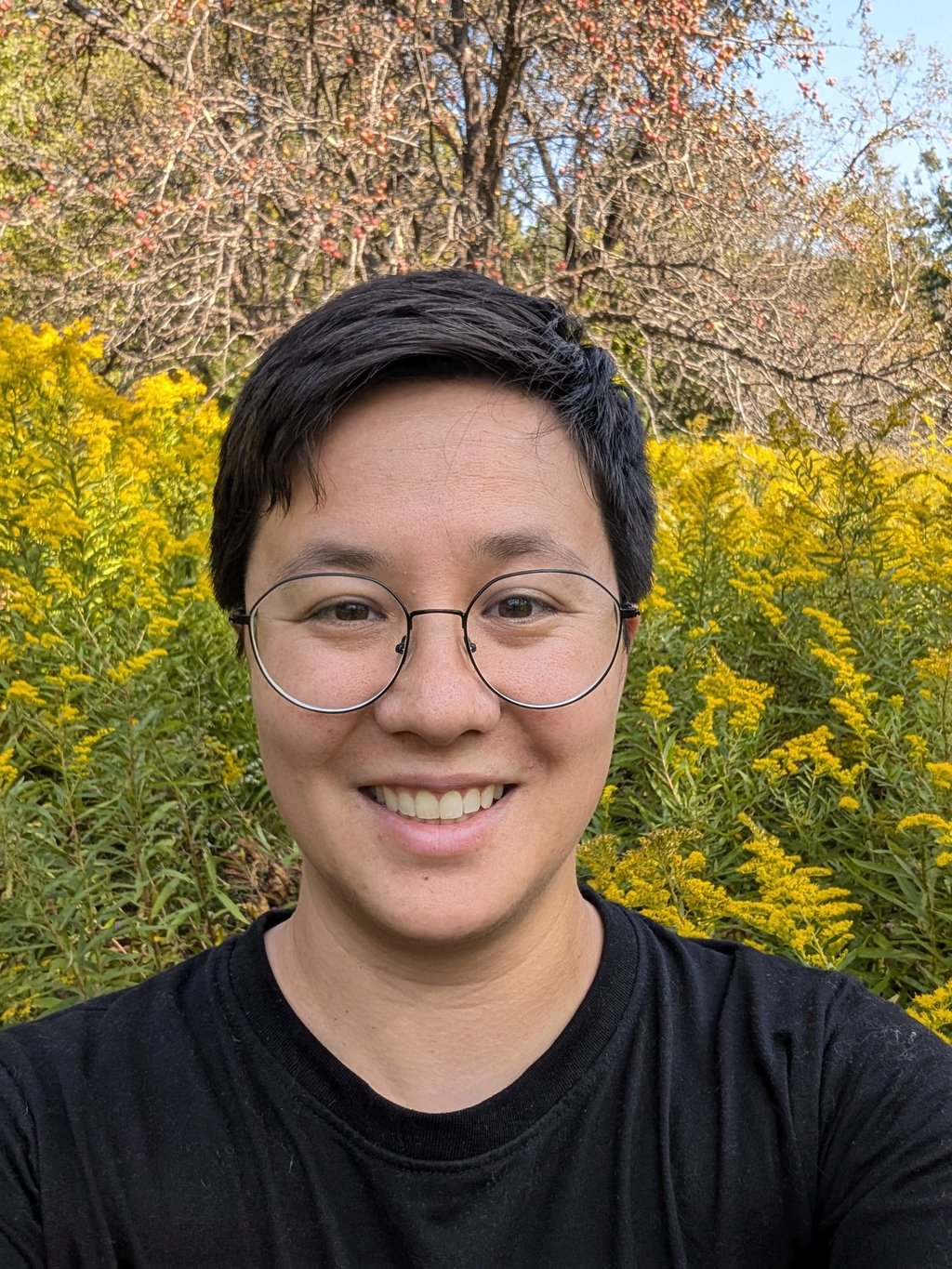 Smiling transgender man with short hair and glasses in front of blooming yellow wildflowers.