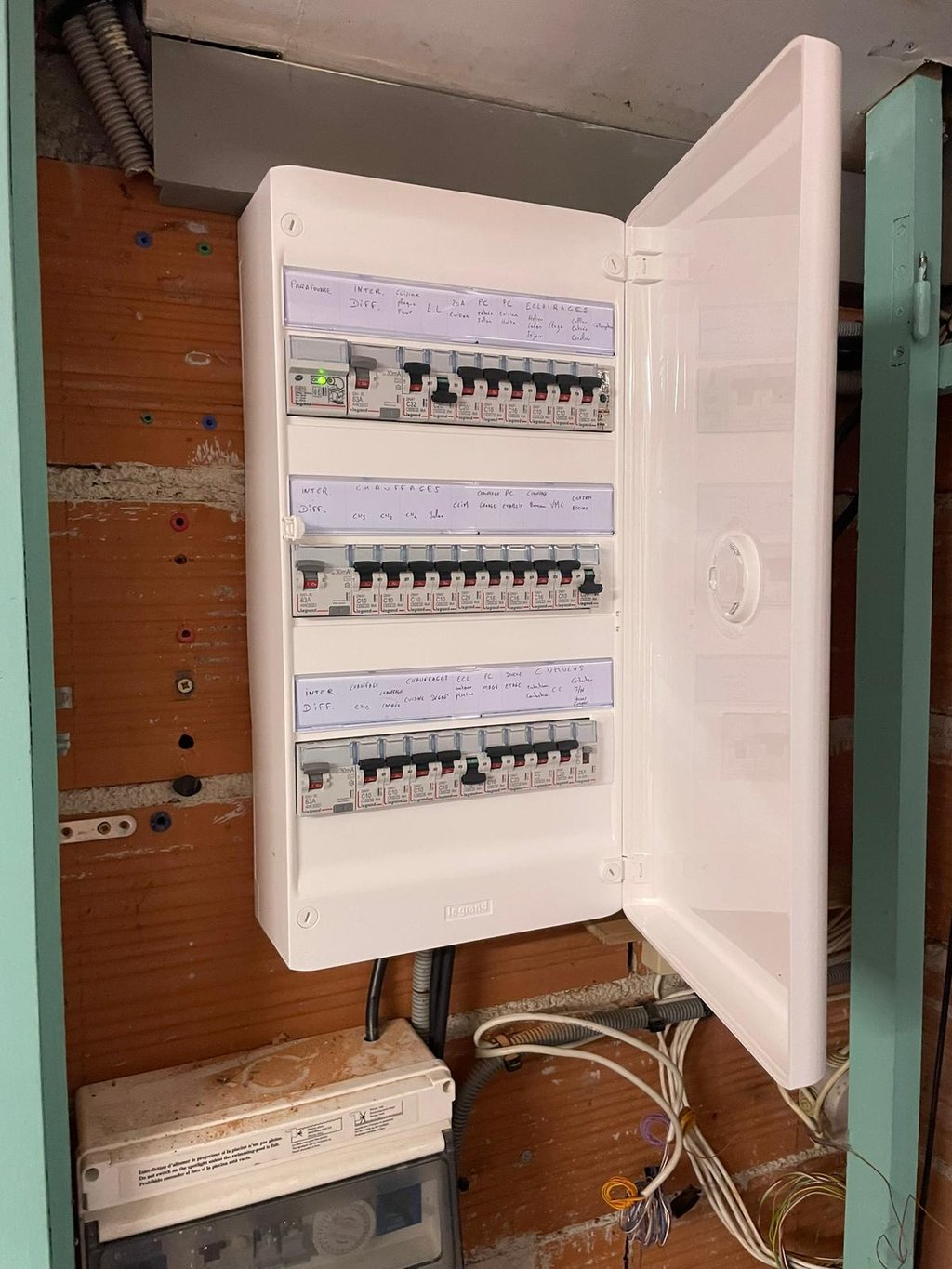 Open white electrical panel board with rows of circuit breakers and handwritten labels on a wall.