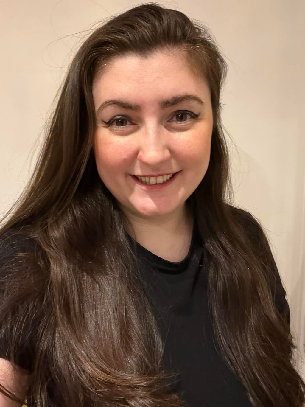 Smiling woman with long dark brown hair and winged eyeliner wearing a black shirt.