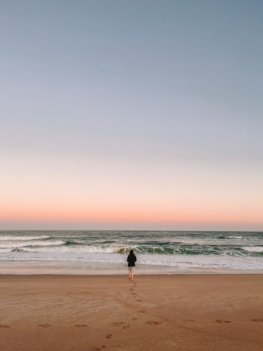 Uma pessoa andando em direção ao mar, de costas, ao por do sol.