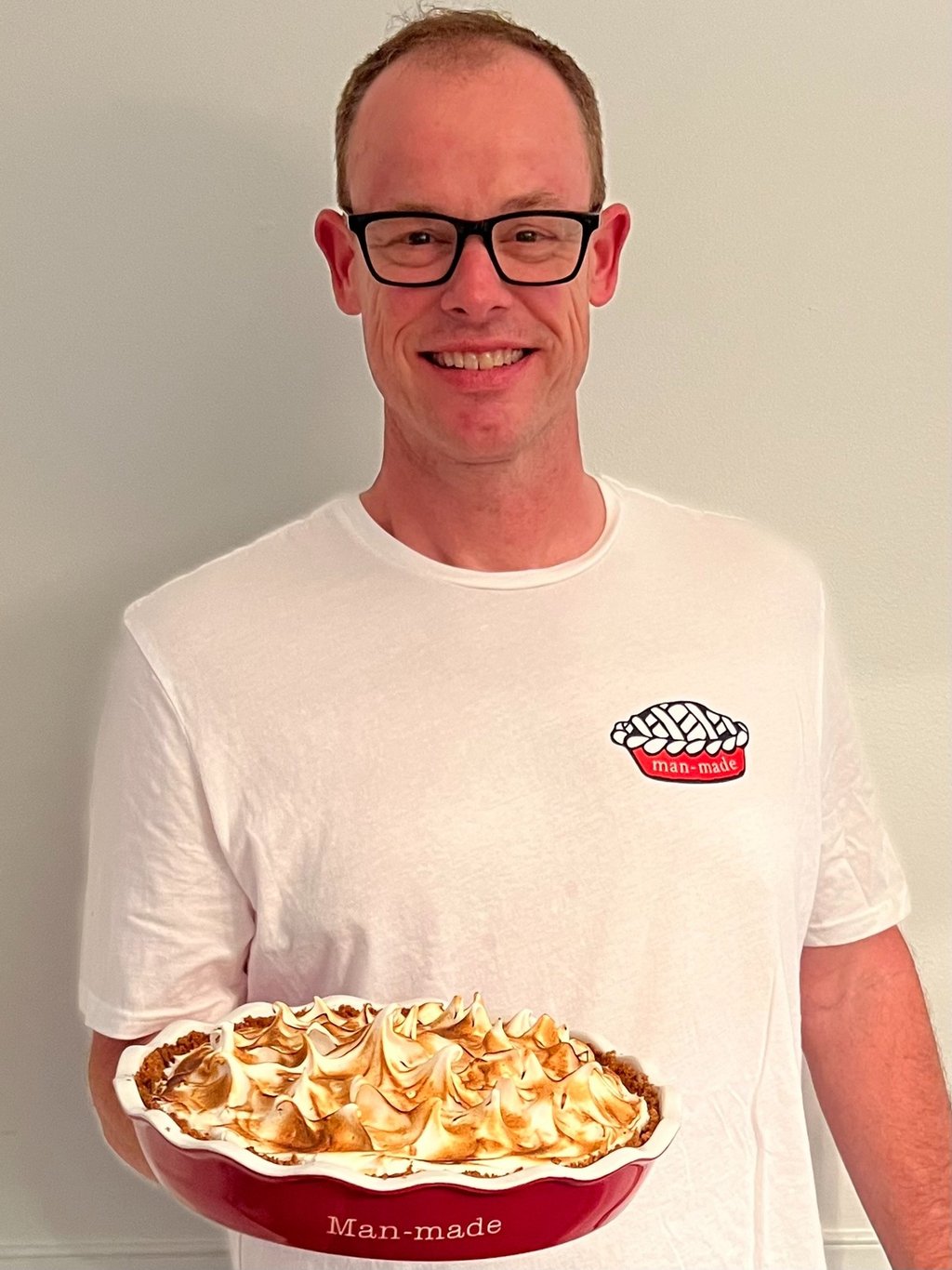 A smiling man wearing glasses holds a freshly baked lemon meringue pie in a red Man-made dish.