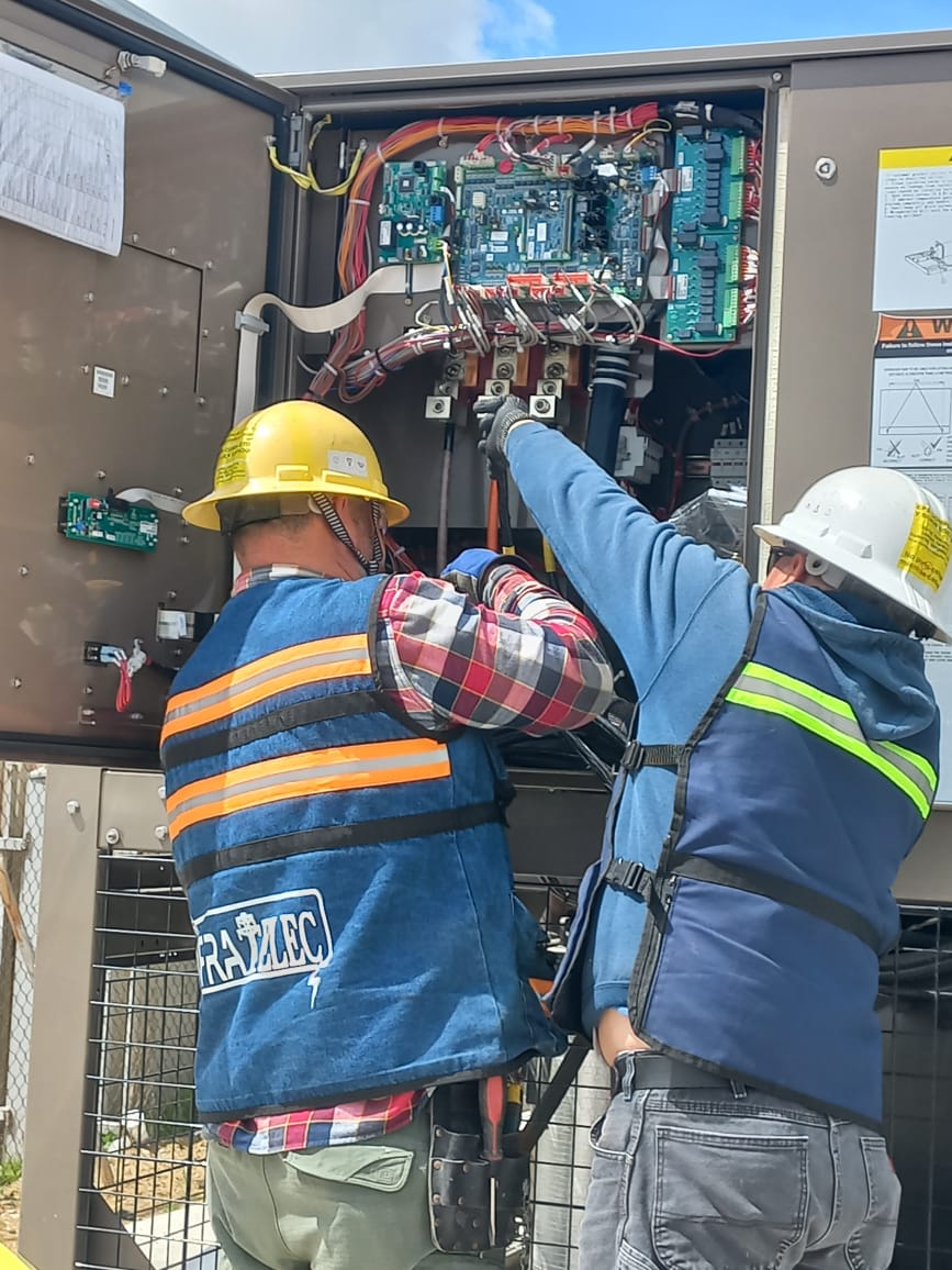 Dos técnicos con casco y chaleco trabajan en panel eléctrico abierto con circuitos.