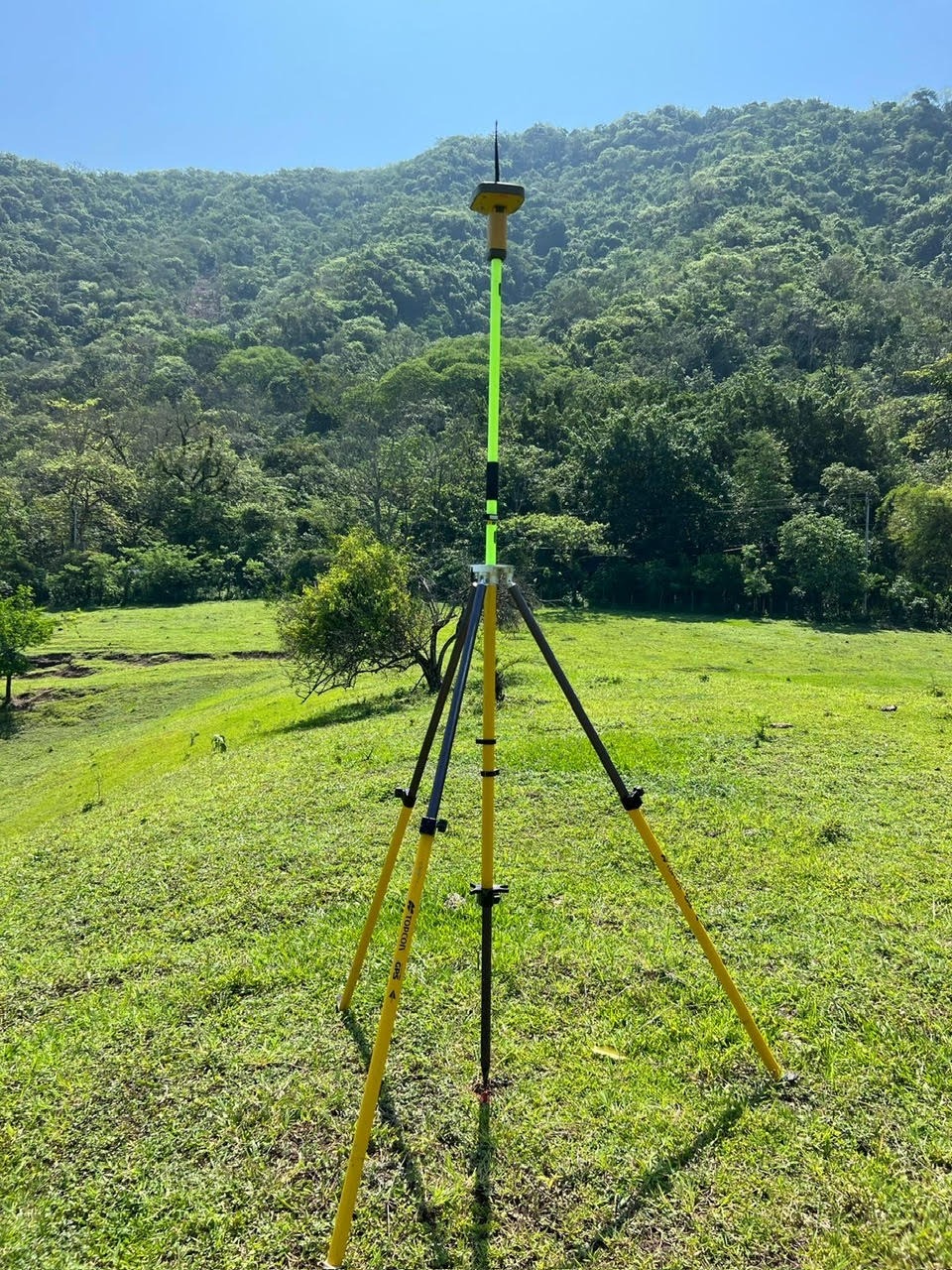 Professional GPS surveying equipment on a tripod in a grassy field with lush green mountains.