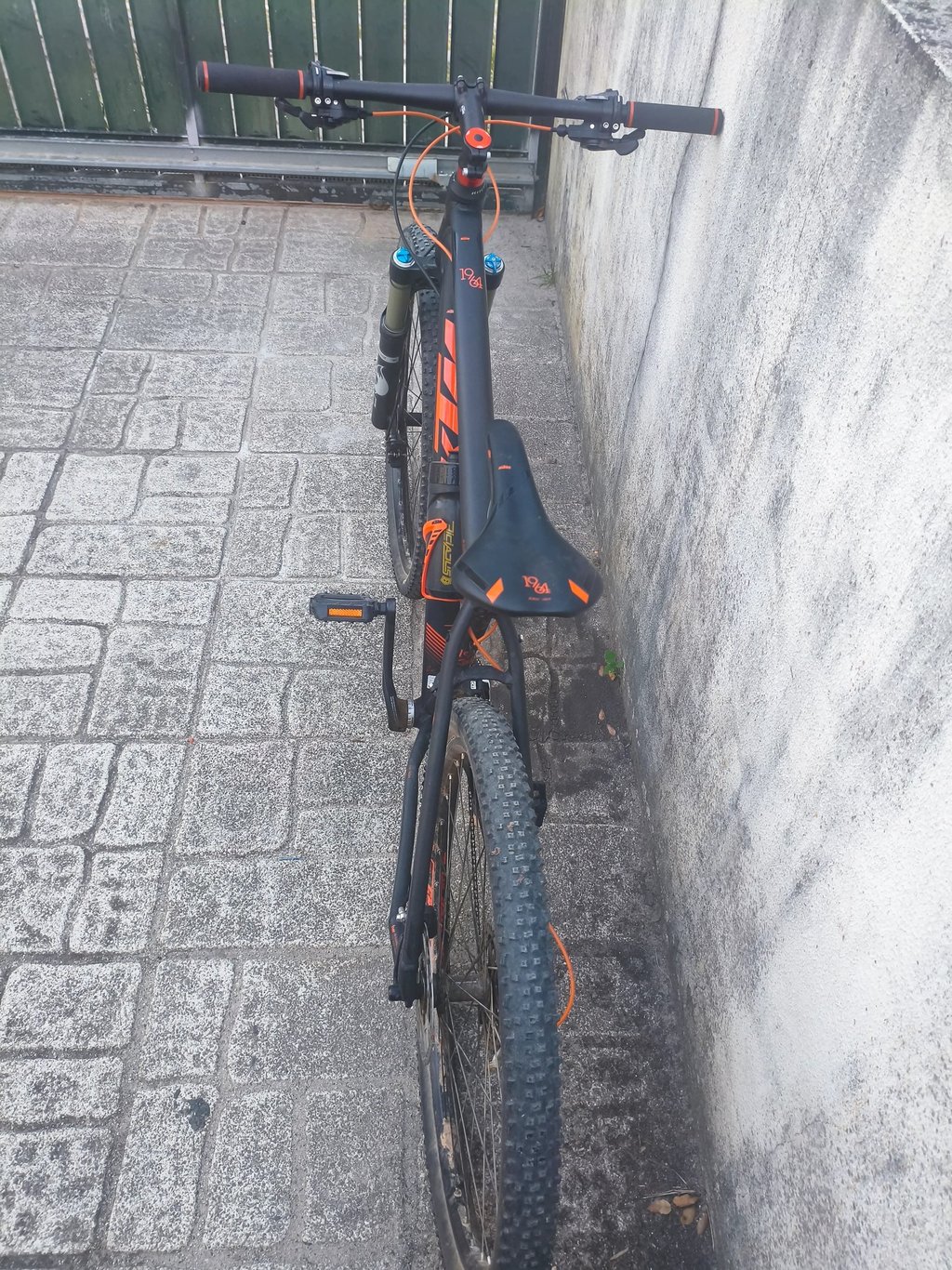 Bicicleta de montanha KTM preta e laranja vista de trás, com selim e pneus largos, apoiada junto a p