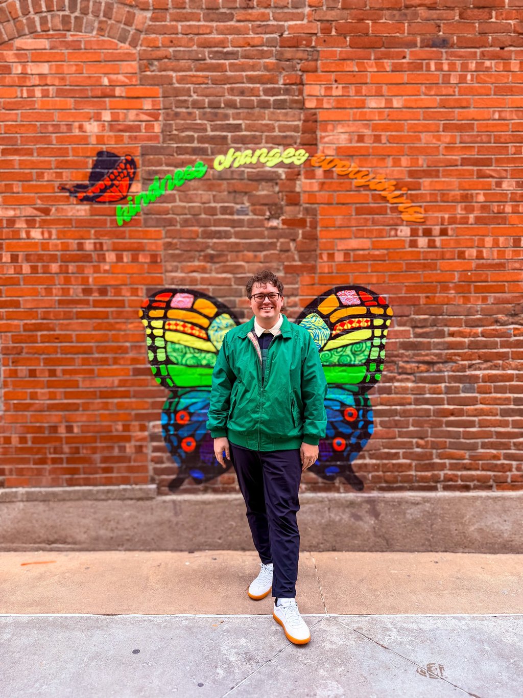 Smiling man in a green jacket posing before a colorful butterfly mural on a red brick wall.