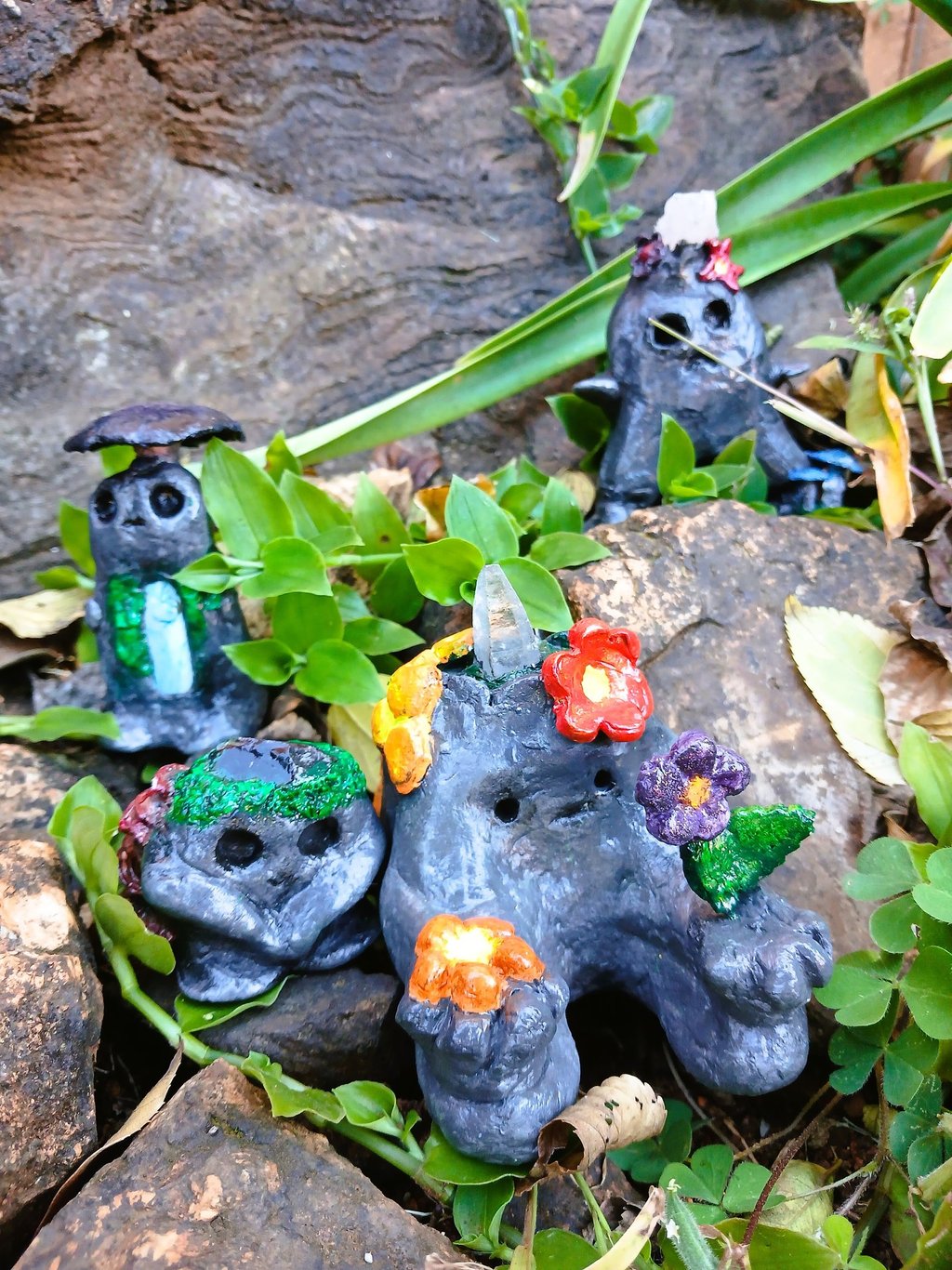 Handmade clay forest spirits with crystals and flowers sitting among garden rocks and green leaves.