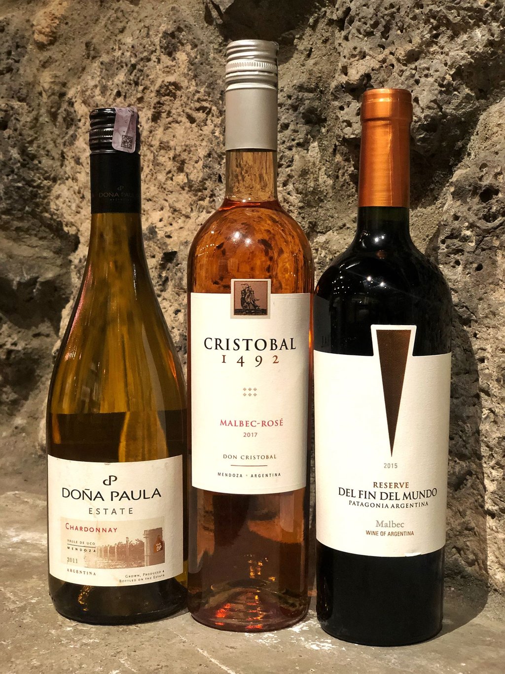 Three bottles of Argentine wine, including Chardonnay, Malbec Rosé, and red Malbec, against a stone wall.