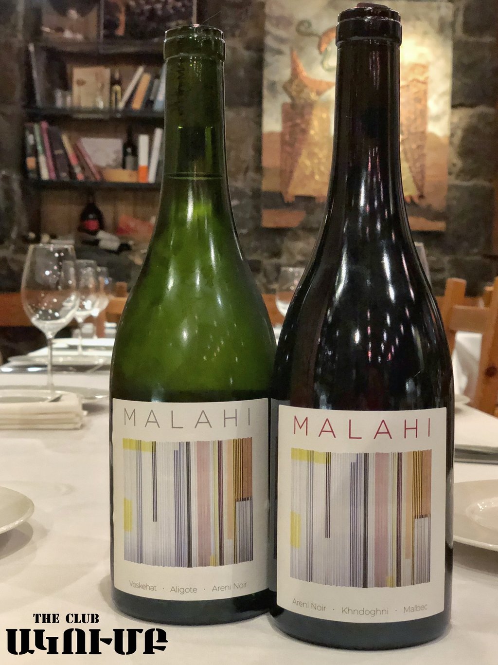 Two bottles of Malahi Armenian wine on a restaurant table with wine glasses and a bookshelf background.