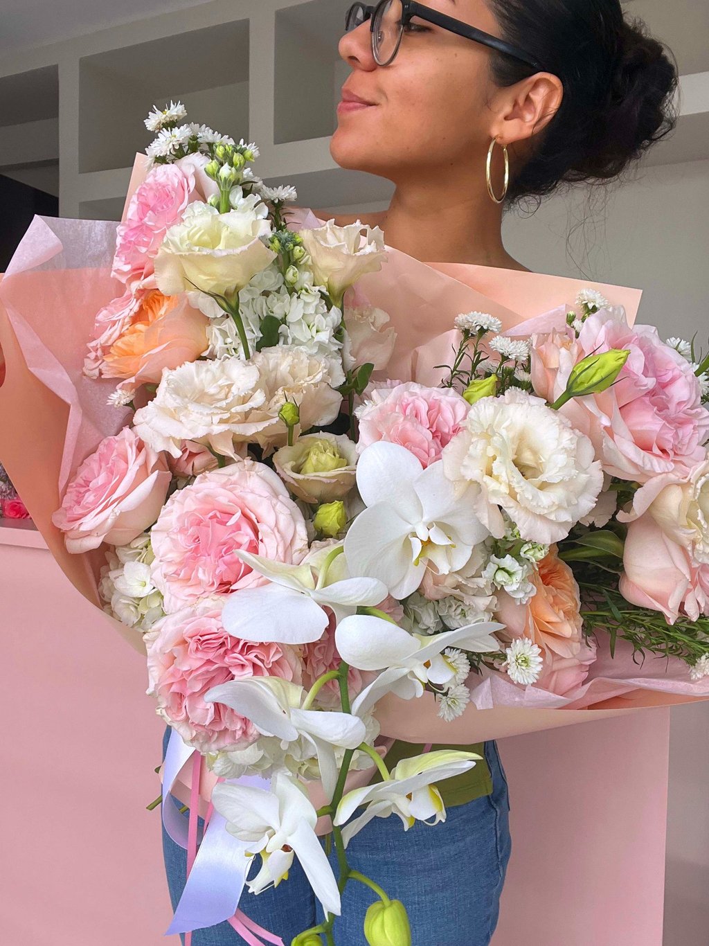 A woman holding a large floral bouquet featuring pink roses, white orchids, and lisianthus flowers.