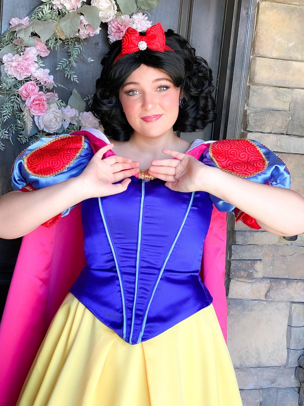 A woman in a Snow White princess cosplay dress with a red bow and floral background.