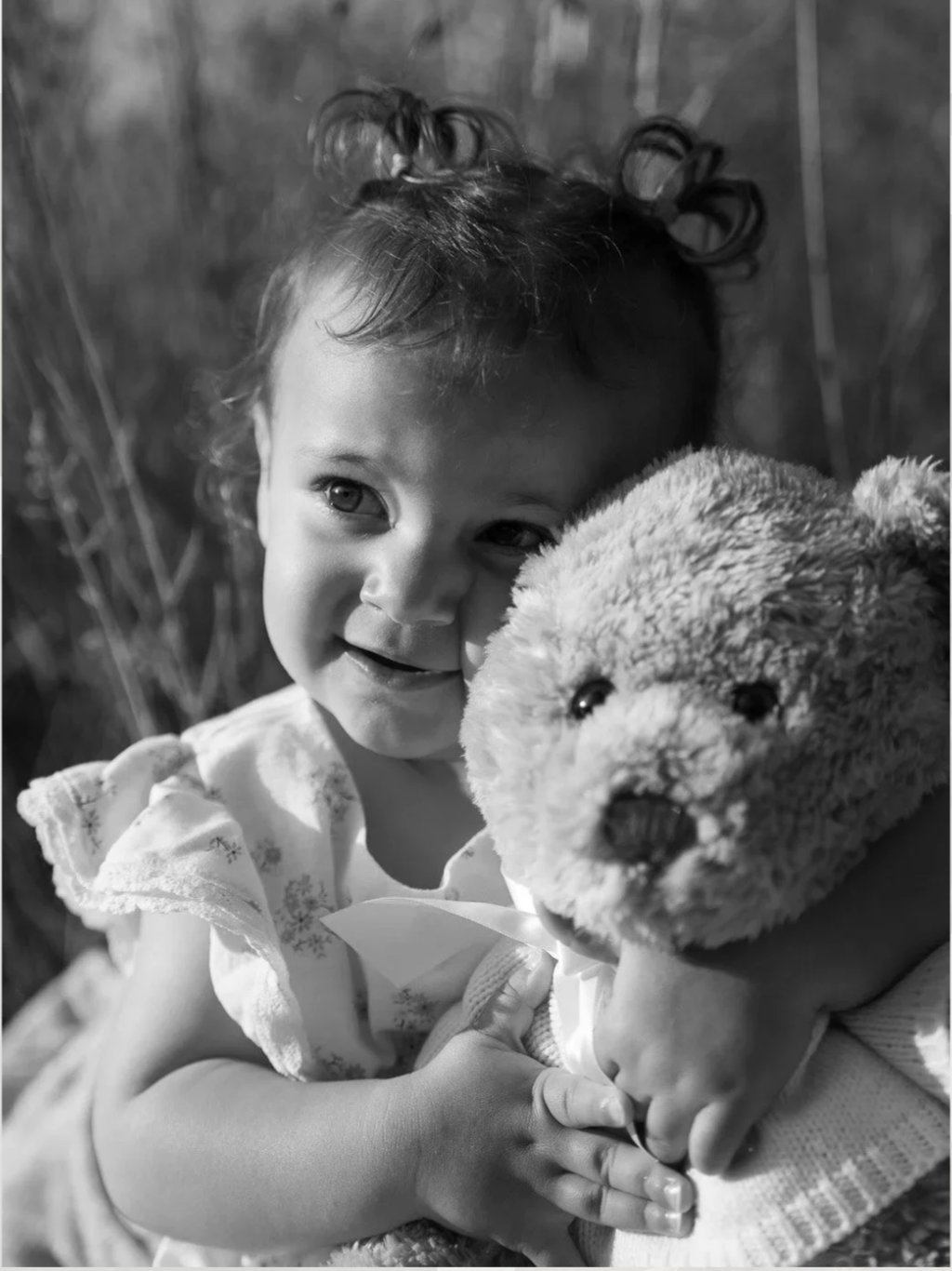 Black and white portrait of a smiling toddler holding a teddy bear during a family photoshoot.