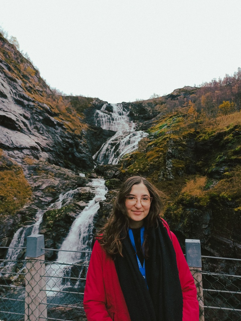 A smiling young woman in a red jacket posing in front of a majestic waterfall in Norway.