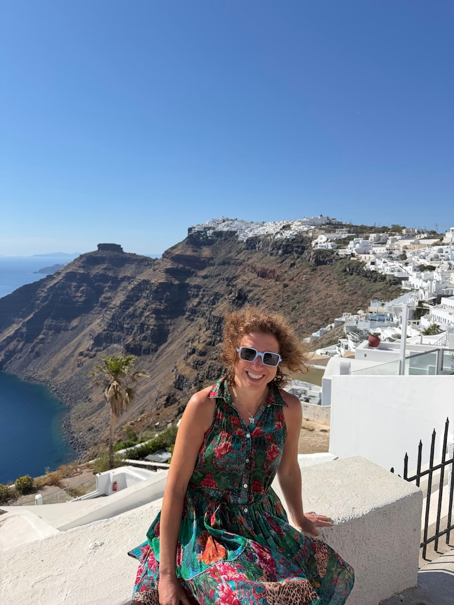 A smiling woman in a floral dress overlooking the Santorini caldera and white buildings of Imerovigli, Greece.