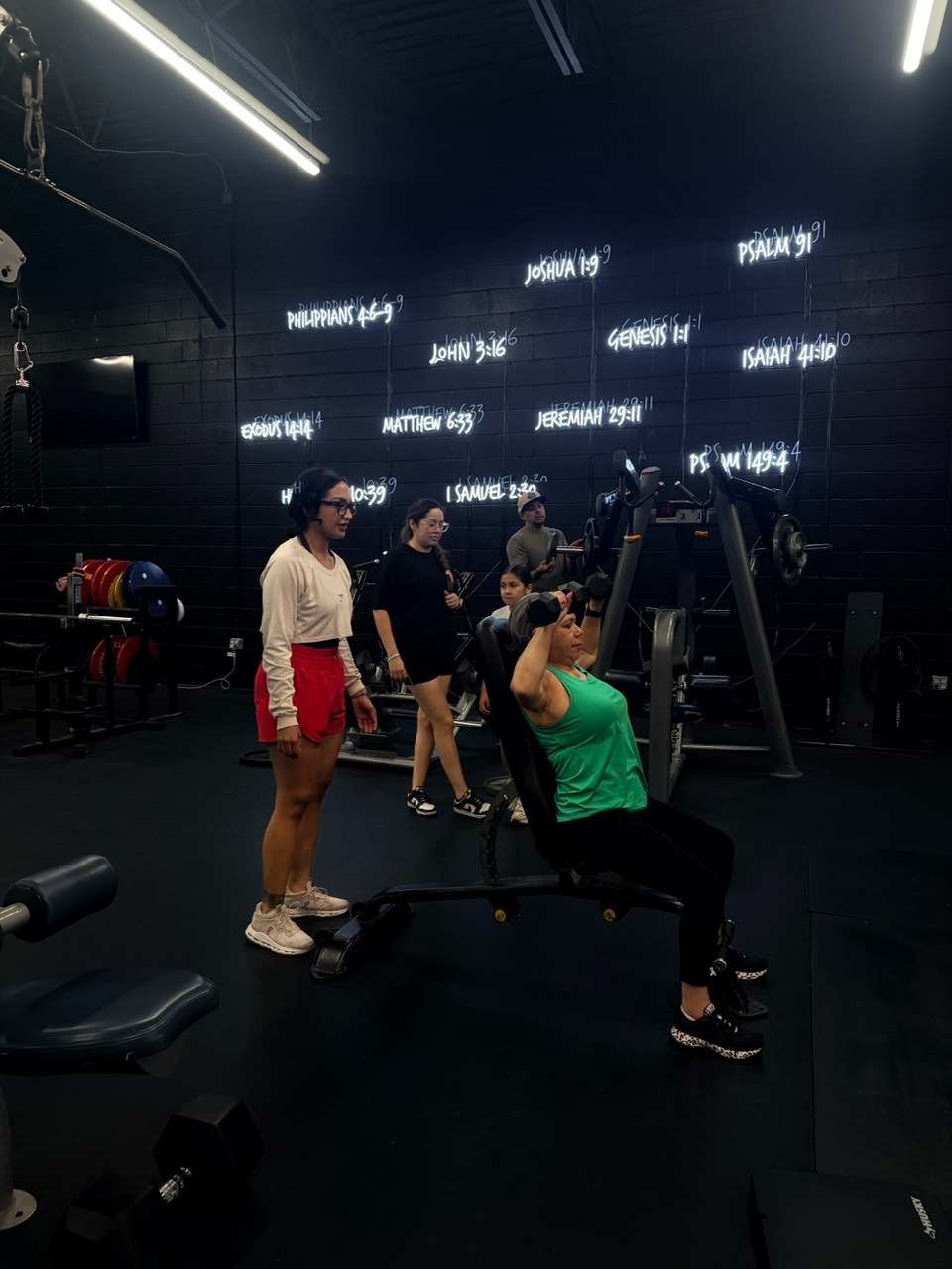 A woman performs a seated dumbbell press at a gym featuring neon Bible verse signs on the wall.