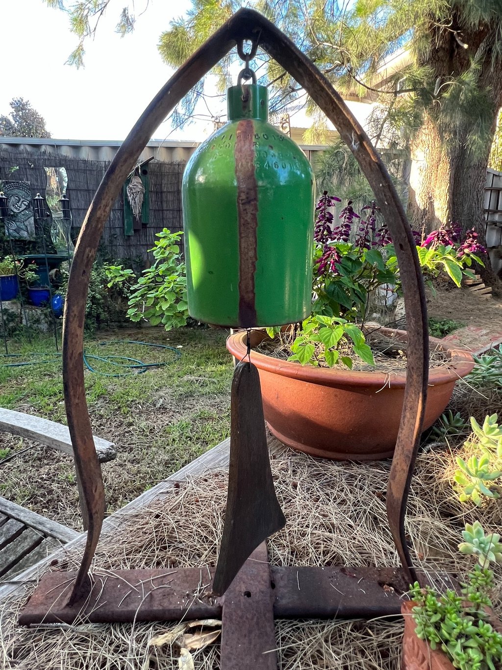 Rustic green metal garden bell wind chime hanging on a weathered iron stand in a backyard garden.