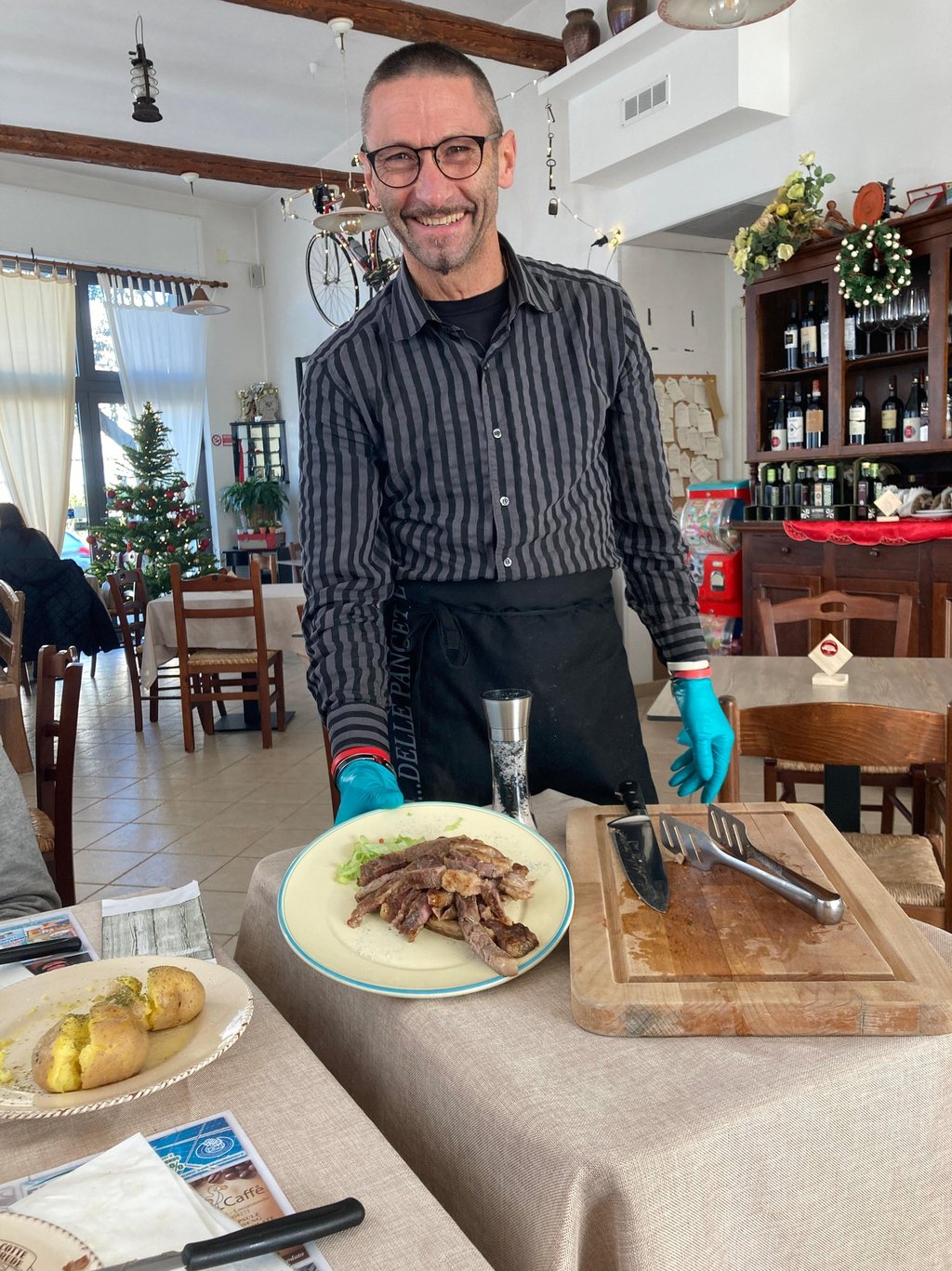 Smiling waiter serving a sliced steak platter in a cozy Italian restaurant with festive decor.