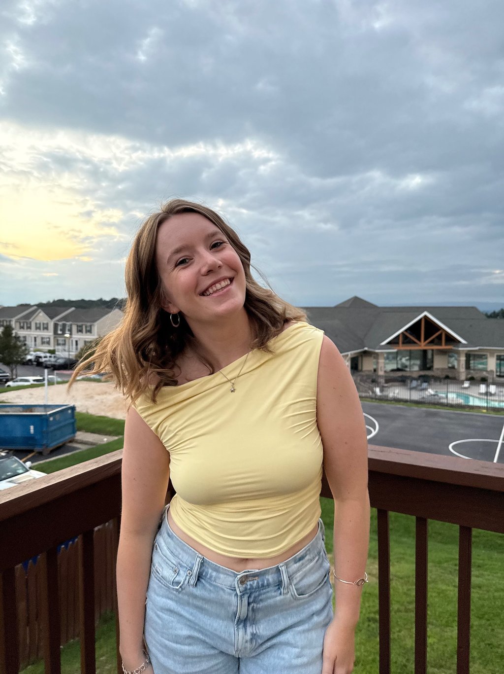 Smiling woman in a yellow top and denim jeans posing on a wooden balcony at sunset.