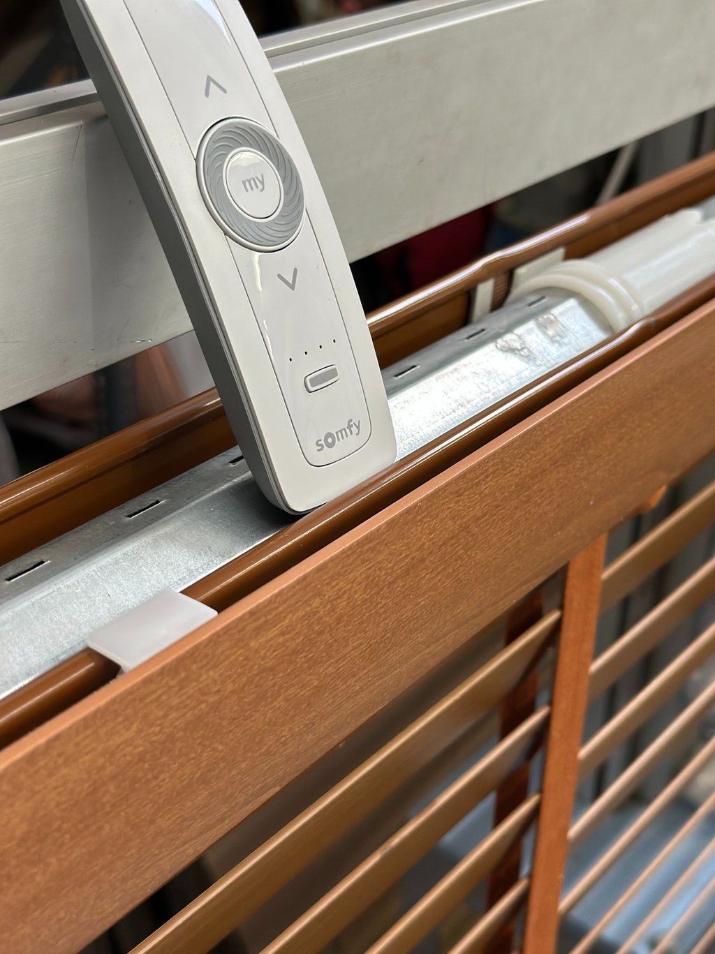 Somfy remote control resting on motorized wooden window blinds with metal hardware.