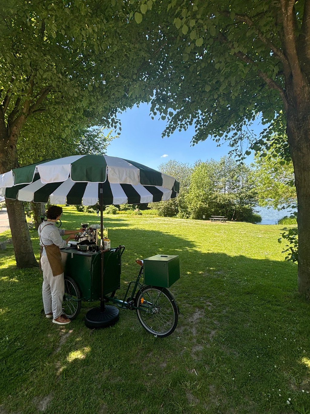 Mobiles dunkelgrünes Kaffeefahrrad mit grün-weißem Schirm am Seeufer.