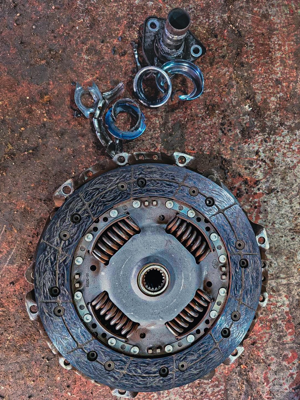 Damaged car clutch plate and broken metal release bearing on a dirty garage floor.