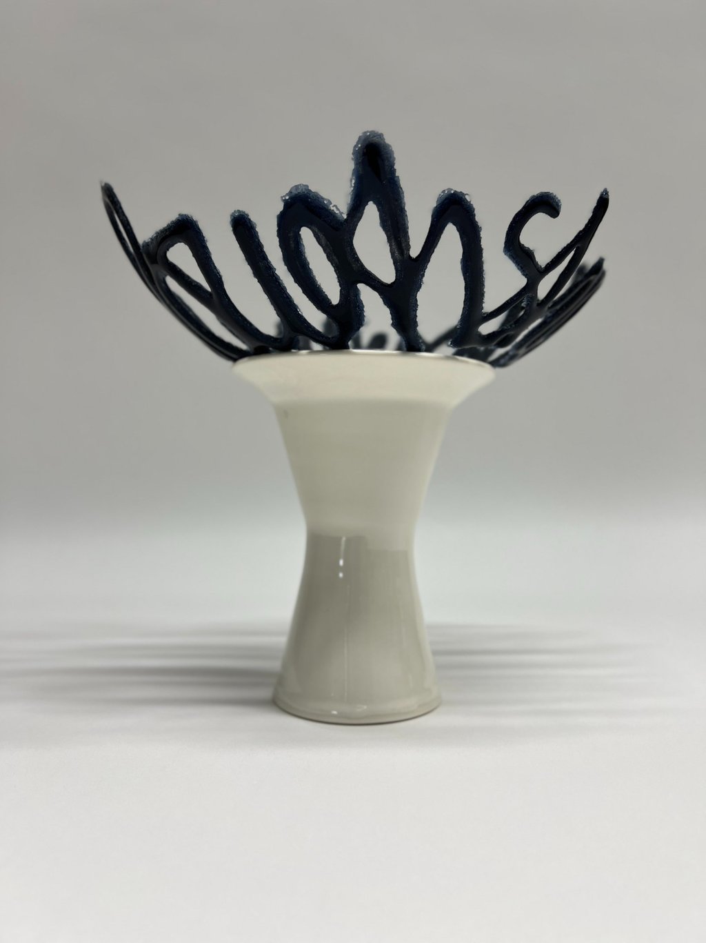 A blue glass bowl sculpture in the shape of a line of text, sitting on a porcelain ceramic pedestal