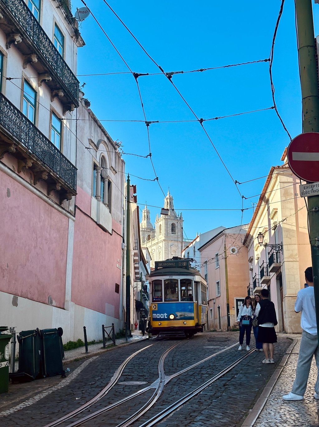 Uma estrada estreira entre as casas portuguesas, com um bondinho ao meio e a catedral ao fundo.