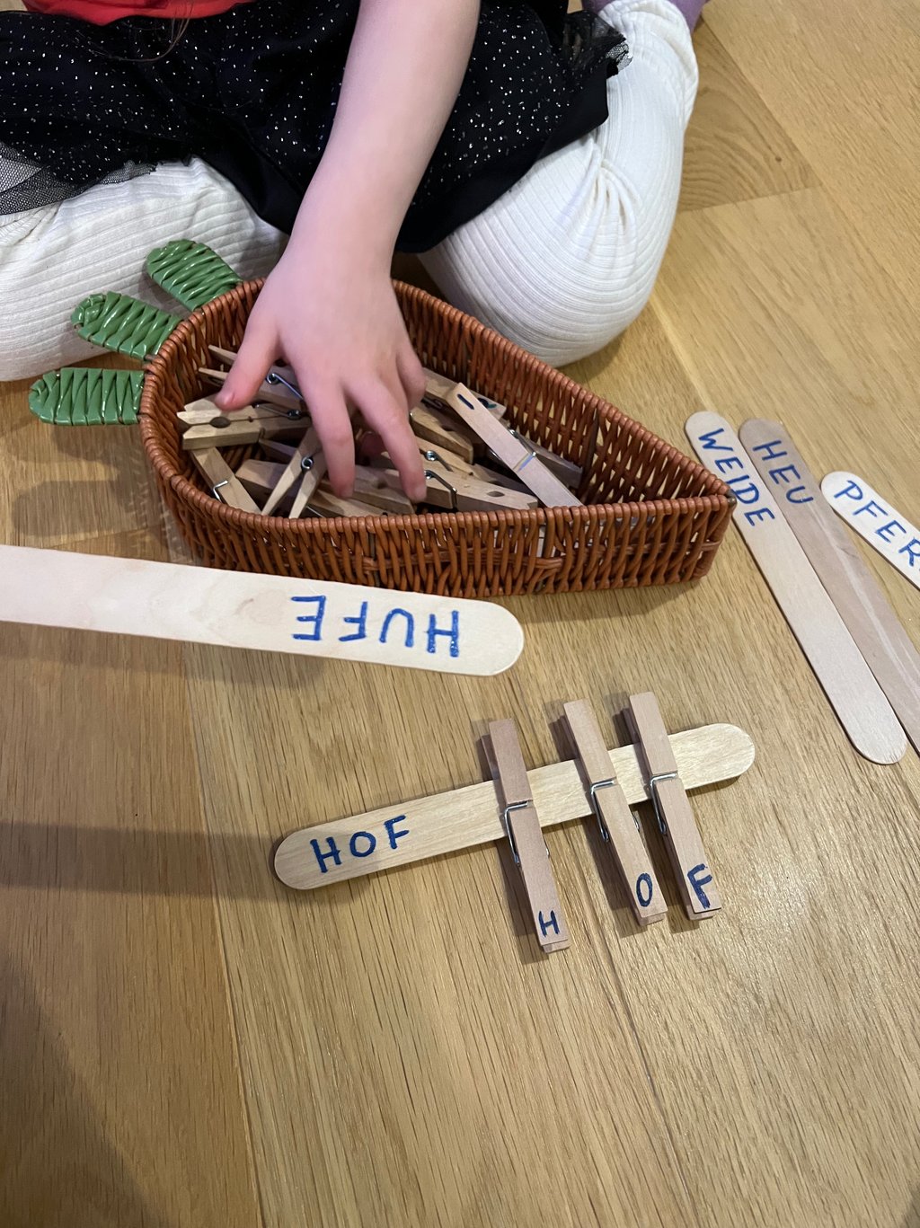 Child using wooden clothespins to match letters on craft sticks for a German word spelling activity.